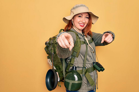 年轻的红发背包客女子在黄色背景上穿着背包和帽子徒步旅行图片