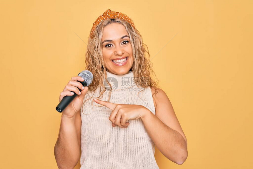 年轻漂亮的金发女歌手在黄色背景上用麦克风用手和指微图片