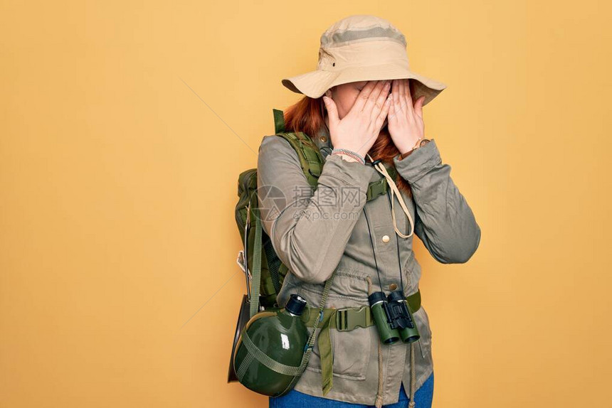 穿着背包和帽子在黄色背景下擦眼的红发年轻穿背心妇女徒步旅行图片