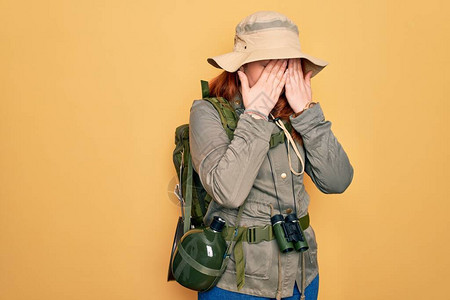 穿着背包和帽子在黄色背景下擦眼的红发年轻穿背心妇女徒步旅行图片