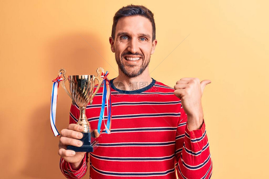 英俊的年轻成功男子赢得奖杯站立在孤立的黄色背景上指着大拇指向那一边笑图片