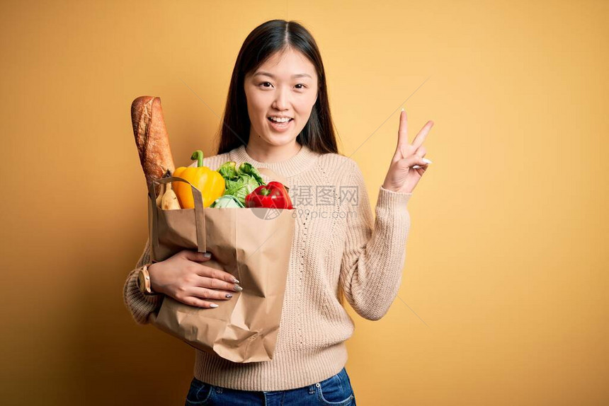 年轻的亚洲女人拿着纸袋满是新鲜健康食品在黄色孤立的背景中微笑地看着摄影机手举起来做胜利标志第二图片