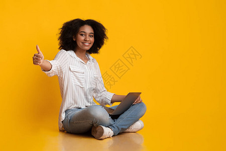 用笔记本电脑和显示缩略图的手势在地上对坐在地板上的非洲女孩微笑图片