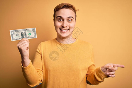 年轻英俊的红发男子拿着一美元钞票在孤立的黄色背景上非常高兴地用手和图片