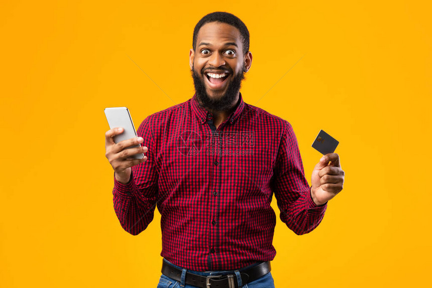 移动购物概念兴奋的非洲裔美国人拿着信用卡和智能手机摆在黄色背景上工作室拍摄快乐的黑图片