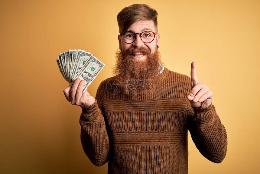 留着胡须的爱尔兰红发男子拿着一堆1美元钞票在黄色背景上图片