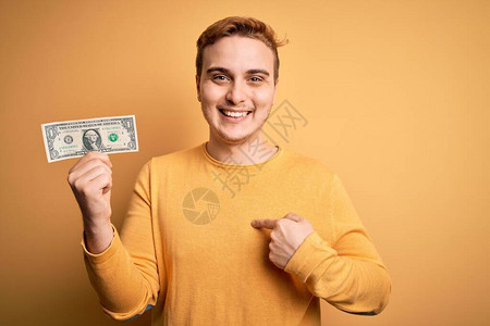 年轻英俊的红头发帅哥在孤立的黄色背景上拿着一美元钞票满脸惊讶地用图片