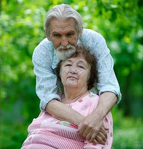 年老的丈夫拥抱坐在椅子上反对绿树叶的老婆图片