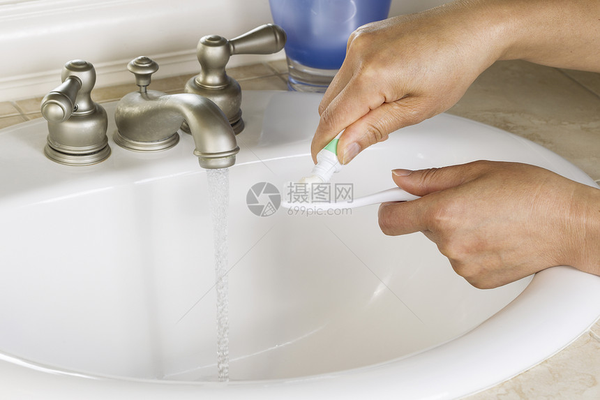 女双手在牙刷上贴牙糊白色浴室水槽蓝色杯子和背面龙头的图片