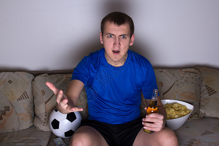 穿着制服坐在沙发上看足球喝着啤酒和薯片的年轻情绪震撼图片