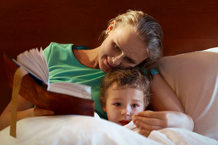年轻母亲读着一本厚的书念给在床上微笑的孩子看图片