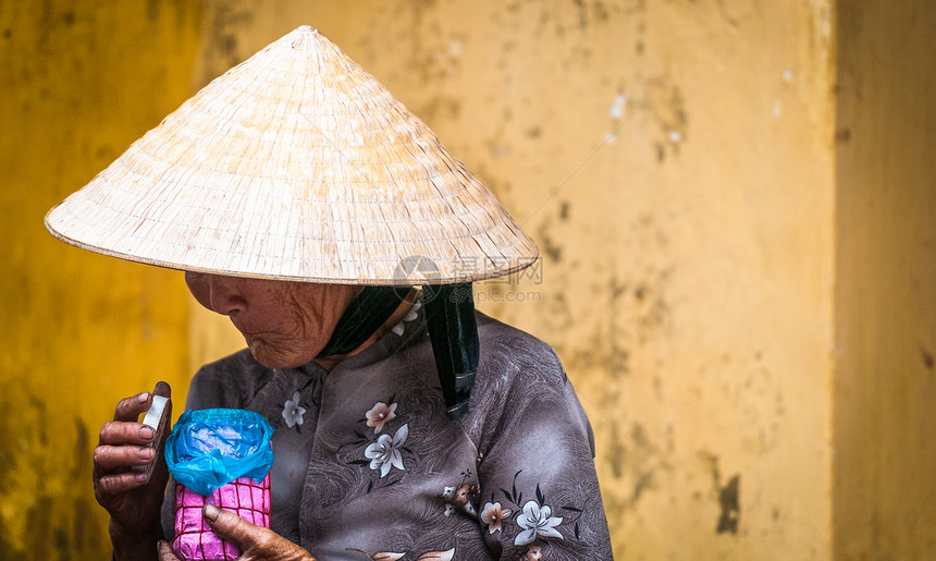 戴着圆锥形帽子和传统亚洲服饰的老可怜的女人拿着粉色和蓝色罐子的越南女士头饰下的皱纹脸站在亚洲越南黄图片