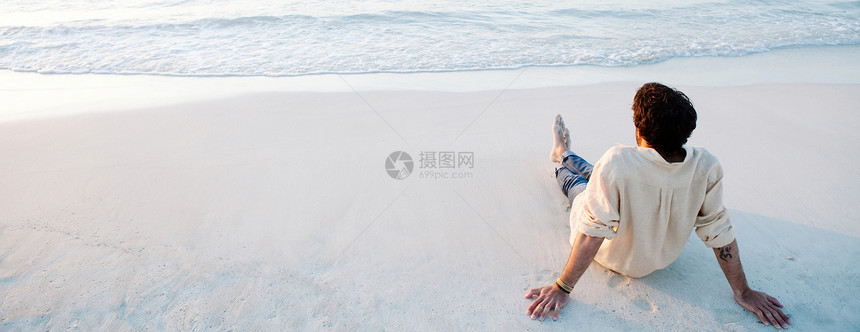 一个年轻人在日落时坐在白色沙滩的海岸上放松的全景格式后视图图片