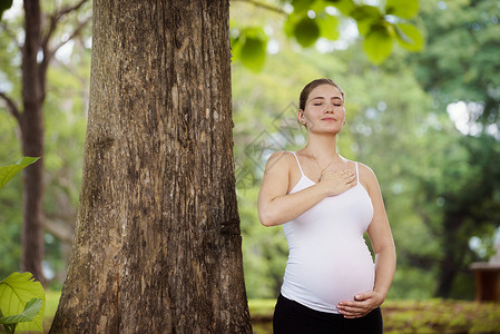 白色美丽的孕妇在树附近的公园里做瑜伽锻炼抚摸肚子图片