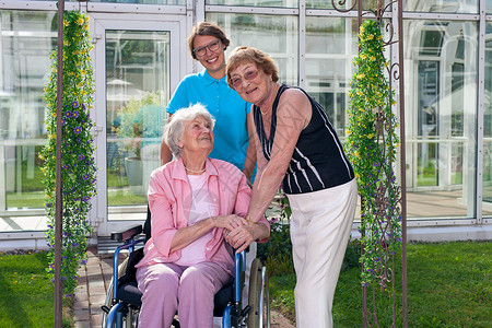 在Glassy绿色花园和玻璃家庭护理大楼坐轮椅的微笑照料顾图片