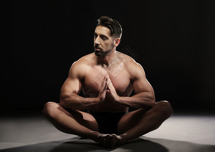 坐在一个瑜伽姿势的无顶部肌肉男的肖像看着黑背景上的摄图片