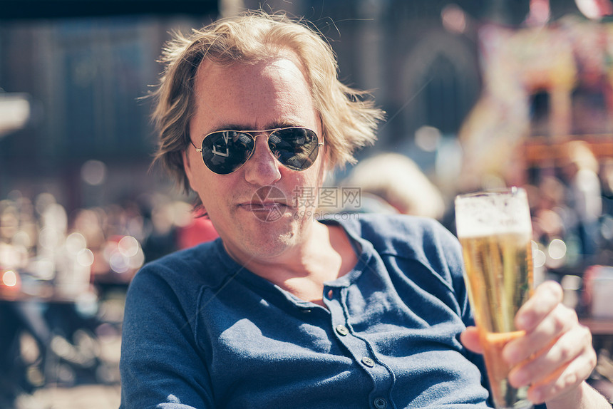 男人在城市广场喝着冰啤酒他坐在太阳镜里享受春光侧图片