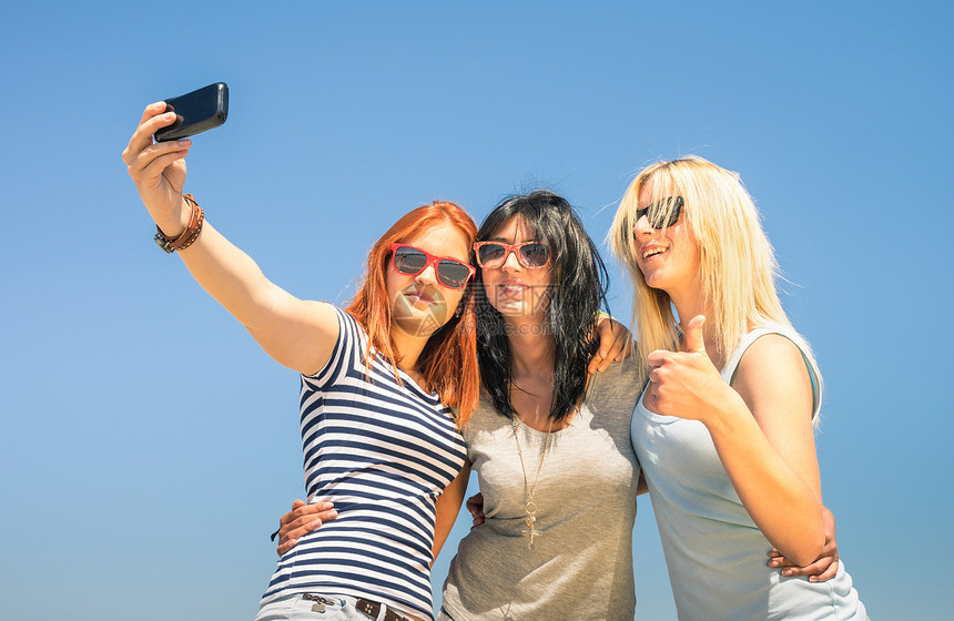 快乐的女朋友在蓝天下自拍友谊夏日概念与新趋势和技术最好的朋友用现代智能手机享受时光温暖阳光明图片