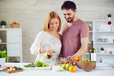 漂亮女人把橄榄油滴到沙拉里和她丈夫在厨图片