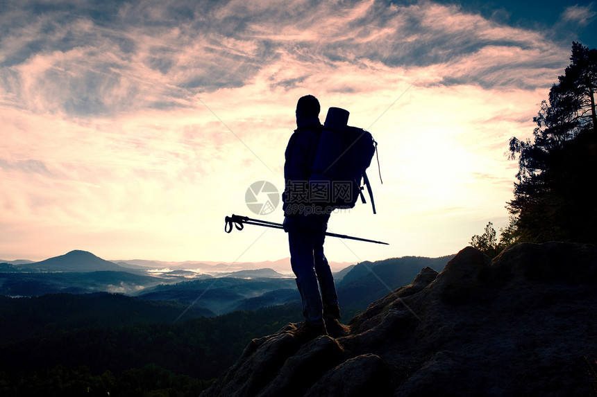 导游提着杆子在路上背着运动背包的徒步旅行者站在迷雾笼罩的山谷上方的岩石观景点上阳光明媚的春天破图片
