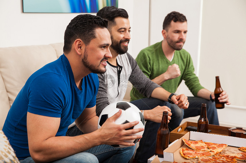 一组三个男朋友一边喝啤酒和吃披萨一边在电视上观看足球比赛的图片