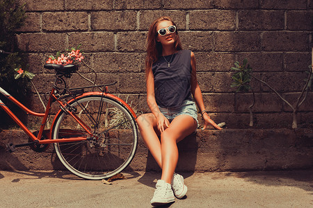 穿着蓝色T恤衫和牛仔短裤的年轻女孩在太阳镜中穿戴着红色自行车图片