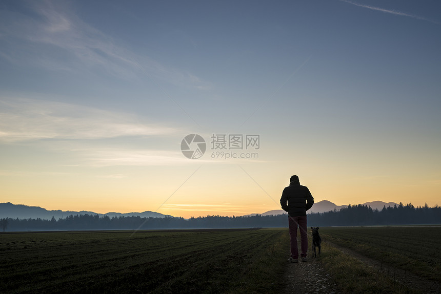 从后面看一个男人在黄昏时分带着他的黑狗走在一条乡间小路上图片