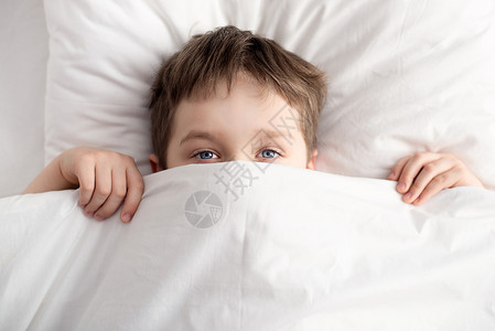 小男孩在床上用白色毯子或床罩遮住脸的顶视图睡觉的男孩图片