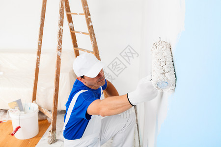 上面的画家用帽子和手套在墙上涂油漆滚木制老旧梯子和背景水桶图片