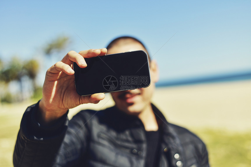 一个穿着胡子的年轻男子在海洋前带着智能手机画图片