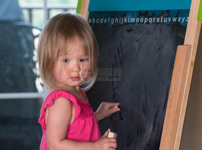 两岁的年轻女孩在黑板上画粉笔并学图片