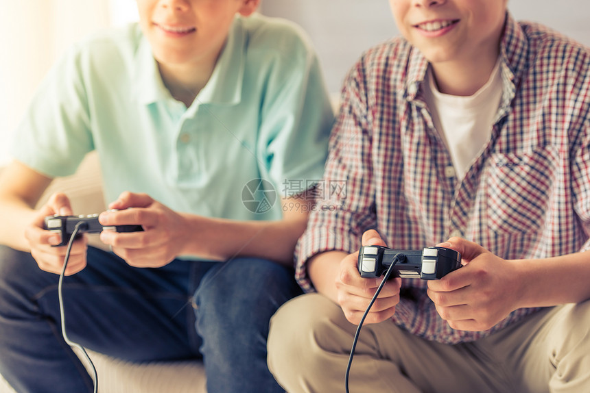 两名有吸引力的少年男孩在家里坐在沙发上玩游戏控制台和笑着微图片