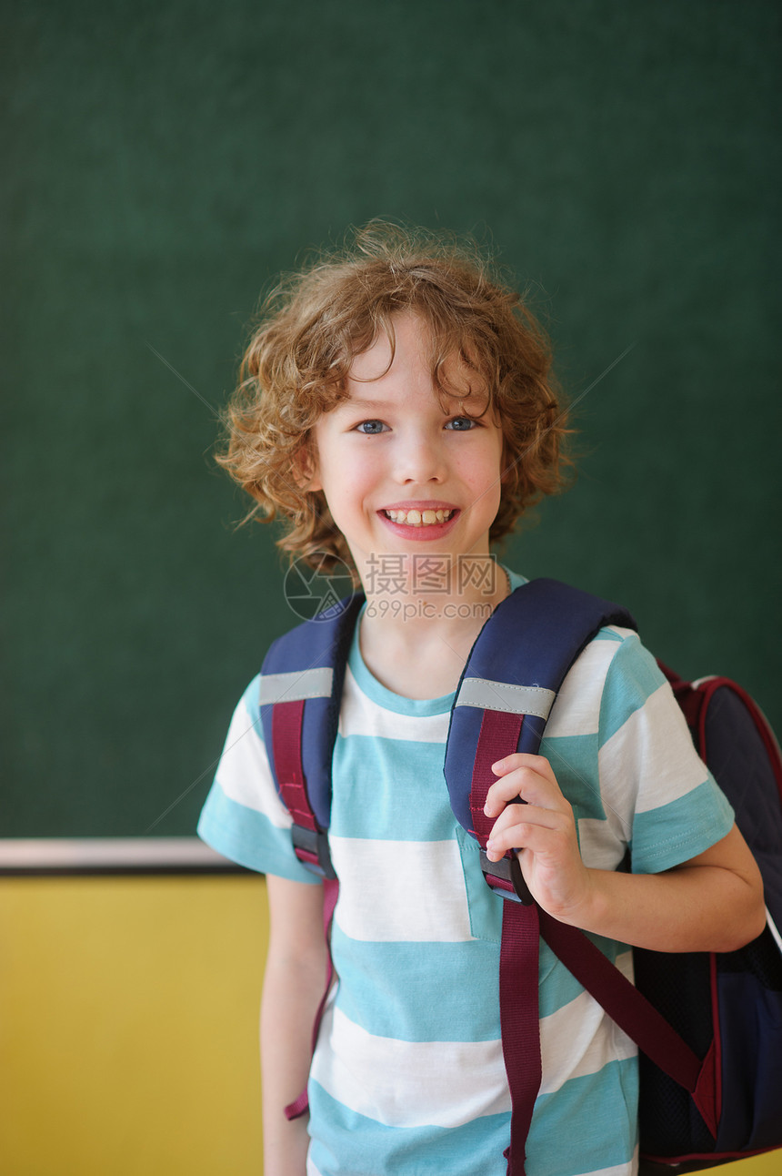 漂亮的小学生站在黑板旁边的班级里在它的肩膀后面是一个书包学校生看着镜头微笑这个男孩有一头的卷发图片