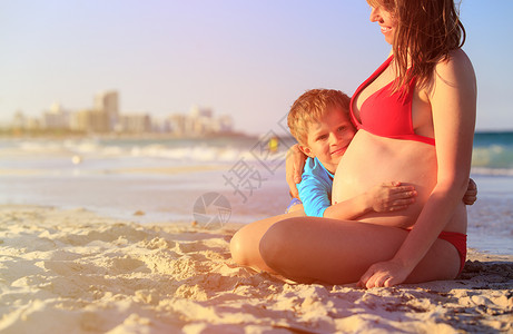 小男孩拥抱怀着孕妇肚子的小男孩在海滩上图片