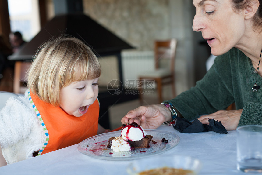 带着橙色围兜的两岁孩子在餐厅里和女人一起吃一块巧克力蛋糕和香草冰淇淋图片