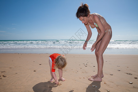 有两岁金发孩子穿橙色冲浪衬衣和比基尼母亲在金沙滩海边采集海壳背景图片
