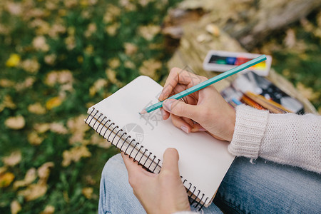 在草地和落叶背景的笔记本中画彩色铅笔关闭在户外笔记本上写字的妇女图片