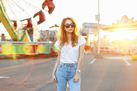 穿着白衬衫的太阳眼镜和古老牛仔裤的漂亮年轻美女在美丽的日落时在游乐图片