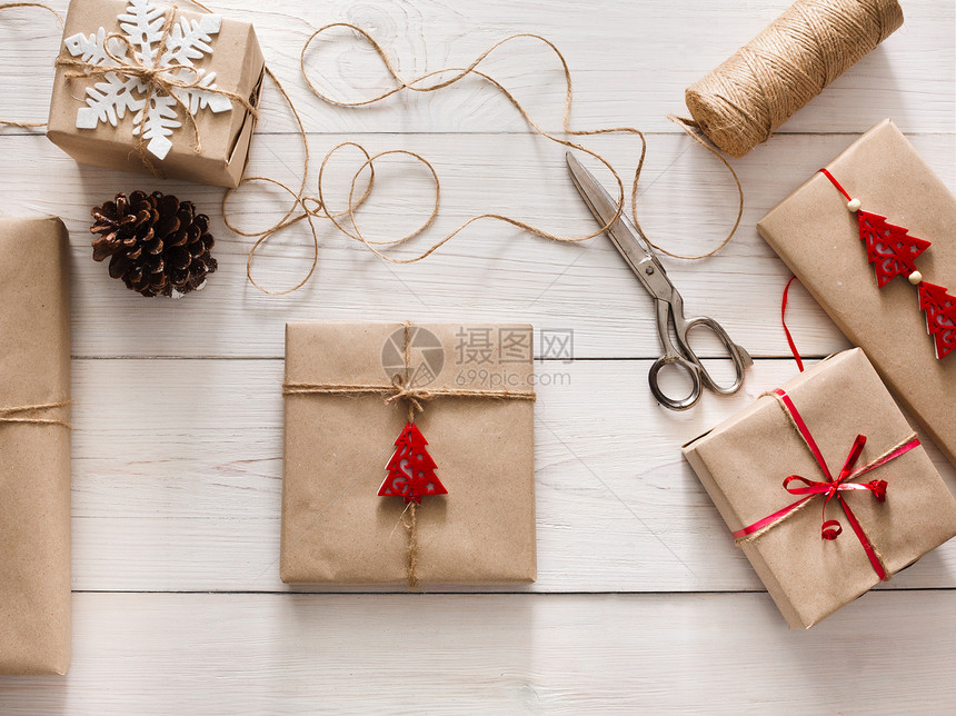 创意爱好用麻线丝带将圣诞假期手工制作的礼物包装在工艺纸中白色木桌上的剪刀和礼品盒图片