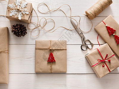 创意爱好用麻线丝带将圣诞假期手工制作的礼物包装在工艺纸中白色木桌上的剪刀和礼品盒背景图片