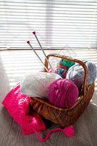 篮子里装着一串毛线和编织针头的球放在有窗户灯光的木制灰色桌子上图片