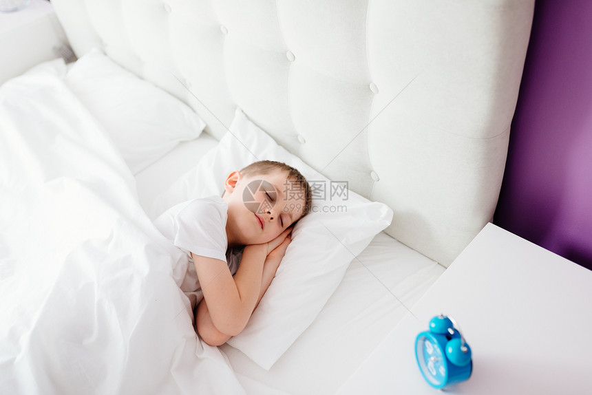 小男孩睡在床上抱着他最喜欢的泰迪熊上面盖着白头巾床边的柜子里有蓝闹图片