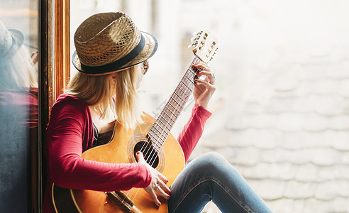年轻快乐的时髦女吉他手穿着休闲时尚服装和夏季时尚帽子坐在家窗前图片