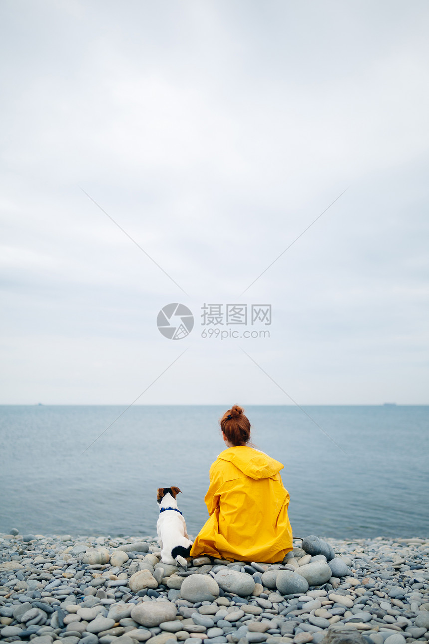 穿着黄色雨衣的女孩背影与狗坐在小石头沙滩图片