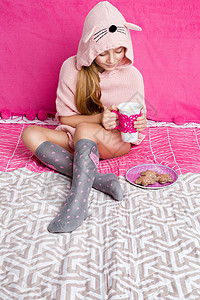 漂亮的小女孩穿着温暖的毛衣兔子耳朵坐在床上喝图片