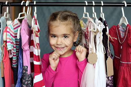可爱的学龄前女孩拥抱在儿童时装店的许多衣服中选择的新可爱连衣裙图片