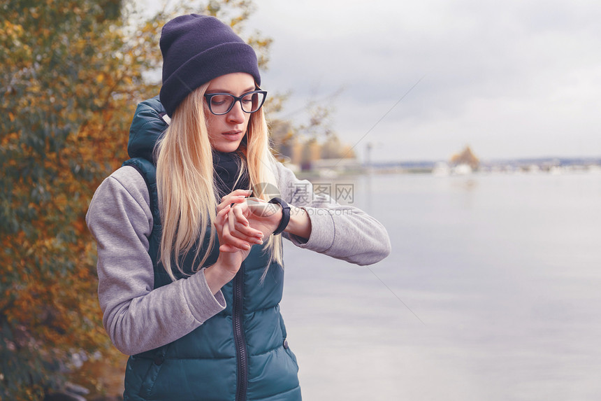 穿着时尚的金发女员调整站在湖岸边的电子手镯计步器秋季风格旅行和健康的图片