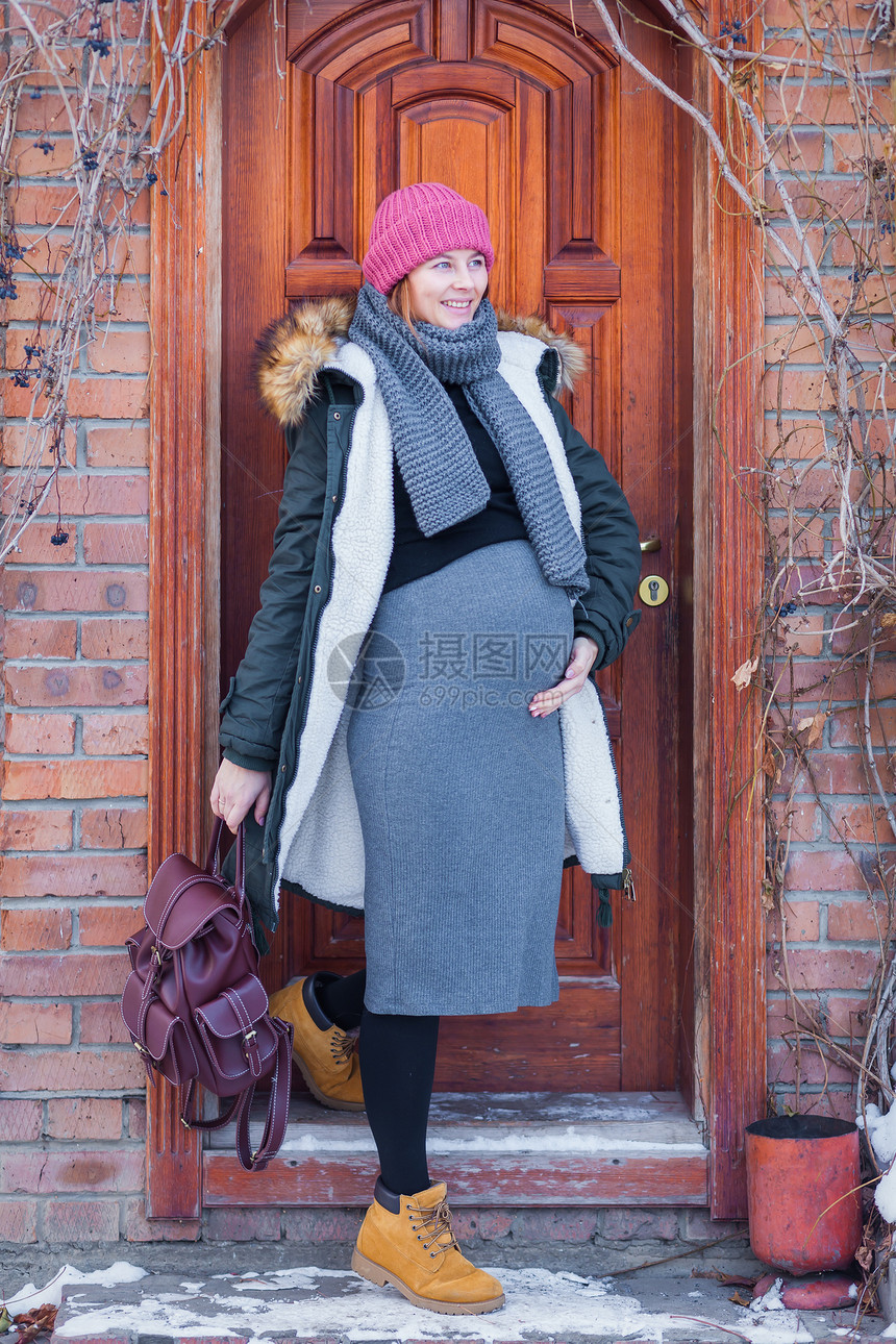 身着针织粉红色帽子灰色礼服温暖外套和背包微笑的孕妇在寒冬走出家外走出去散步图片