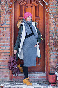 身着针织粉红色帽子灰色礼服温暖外套和背包微笑的孕妇在寒冬走出家外走出去散步背景图片