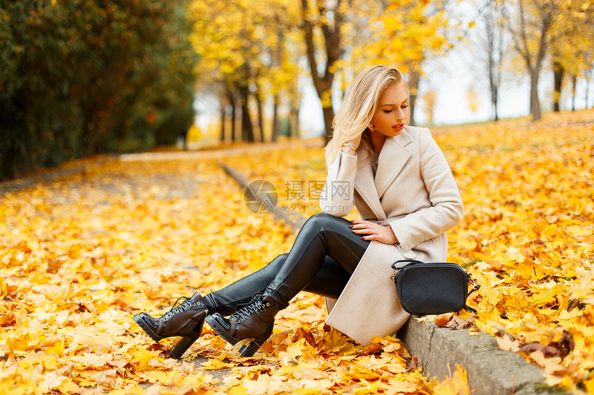 穿着时髦外套穿着时髦鞋子和手提包的漂亮时尚年轻金发女人坐在公园里图片
